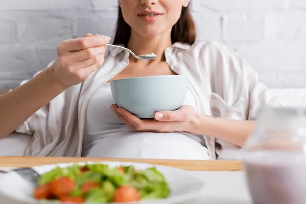 Vue recadrée d'une femme enceinte heureuse mangeant des flocons d'avoine près d'un plateau avec salade — Photo de stock