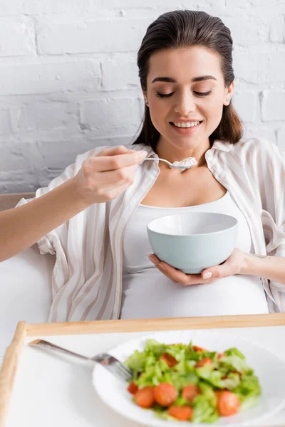 Happy pregnant woman eating oatmeal near tray with salad — Stock Photo