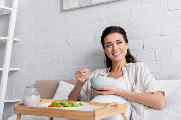 Happy pregnant woman holding bowl with oatmeal near tray with salad — Stock Photo