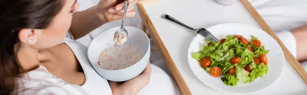 Vista de ángulo alto de la mujer embarazada sosteniendo tazón con harina de avena cerca de bandeja con ensalada, pancarta - foto de stock