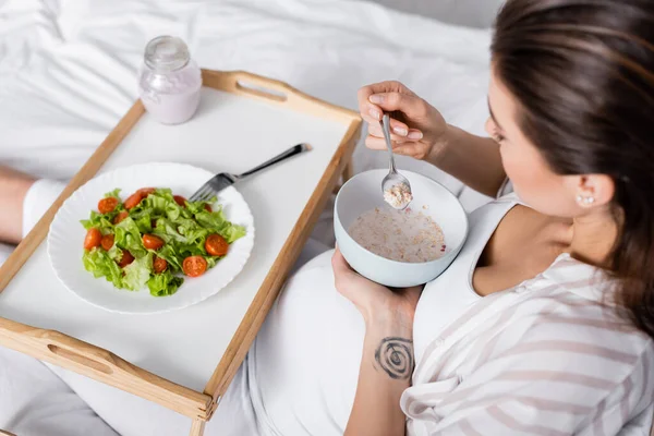 Высокий угол обзора беременной женщины, держащей миску с овсянкой возле подноса с салатом — стоковое фото