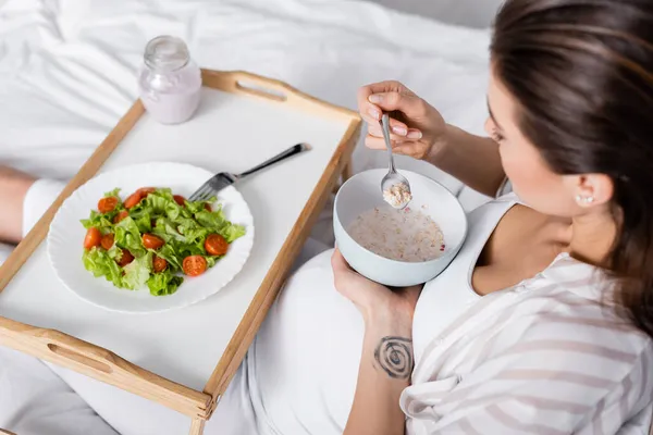 Vista de ángulo alto de la mujer embarazada sosteniendo tazón con harina de avena cerca de bandeja con ensalada - foto de stock