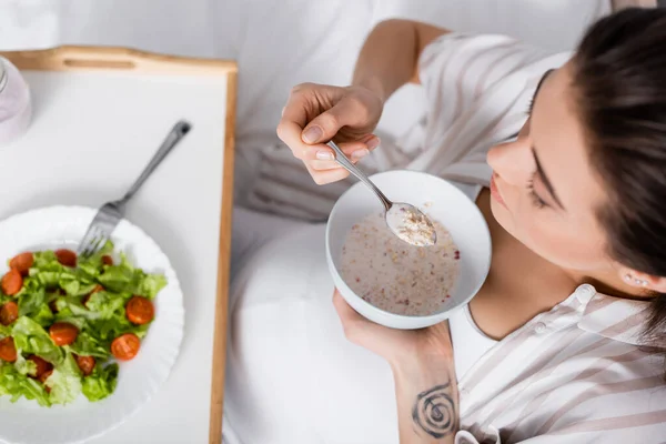 Top view of pregnant woman holding bowl with oatmeal near tray with salad — Stock Photo