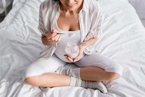 Vista recortada de la mujer embarazada feliz sosteniendo frasco de vidrio con yogur mientras está sentado en la cama - foto de stock