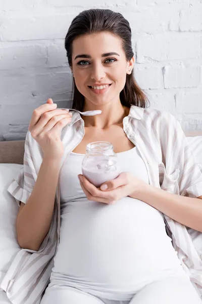 Gaie femme enceinte tenant pot en verre avec du yaourt tout en étant assis sur le lit — Photo de stock