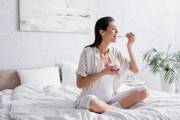 Веселая беременная женщина держит стеклянную банку и ест йогурт, сидя на кровати — стоковое фото