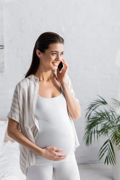 Mujer embarazada sonriente hablando en el teléfono inteligente en el dormitorio - foto de stock