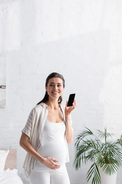 Mujer embarazada sonriente sosteniendo teléfono inteligente en el dormitorio - foto de stock