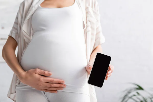 Vista recortada de la joven embarazada sosteniendo teléfono inteligente con pantalla en blanco - foto de stock