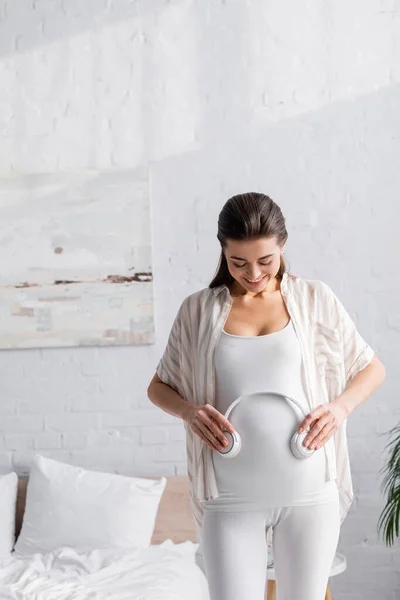 Mujer embarazada feliz sosteniendo auriculares inalámbricos cerca del vientre - foto de stock