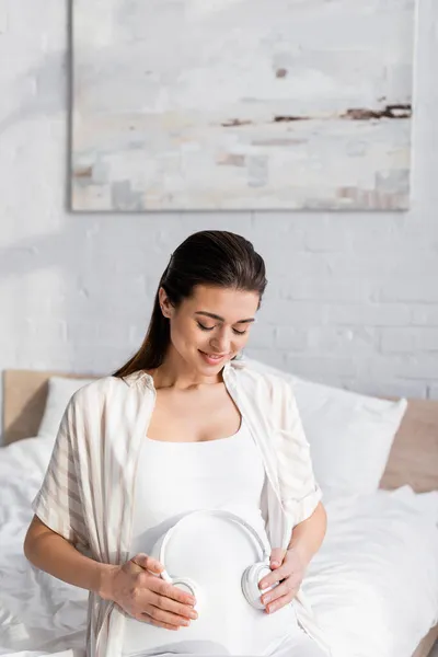 Mujer embarazada complacida sosteniendo auriculares inalámbricos cerca del vientre - foto de stock
