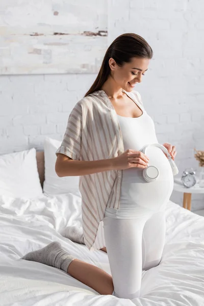 Mujer embarazada alegre sosteniendo auriculares inalámbricos cerca del vientre - foto de stock