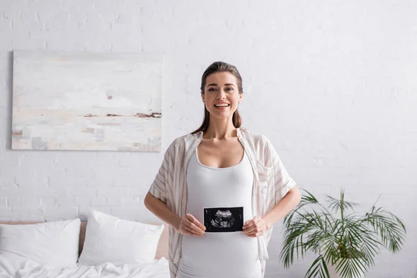 Mujer sonriente y embarazada sosteniendo ultrasonido escanear en el dormitorio - foto de stock