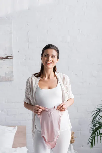Pleased pregnant woman holding pink baby romper in bedroom — Stock Photo
