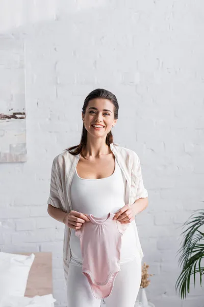 Mujer embarazada complacida sosteniendo rosa bebé mameluco en dormitorio - foto de stock