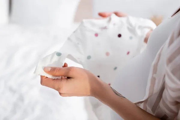 Vista recortada de mujer embarazada tatuada sosteniendo bebé mameluco en el dormitorio - foto de stock