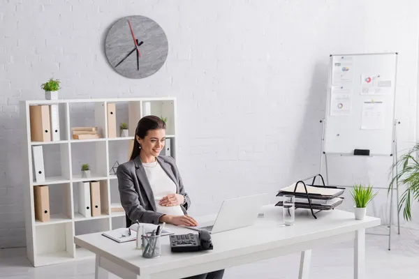 Щаслива вагітна бізнес-леді дивиться на ноутбук в офісі — стокове фото