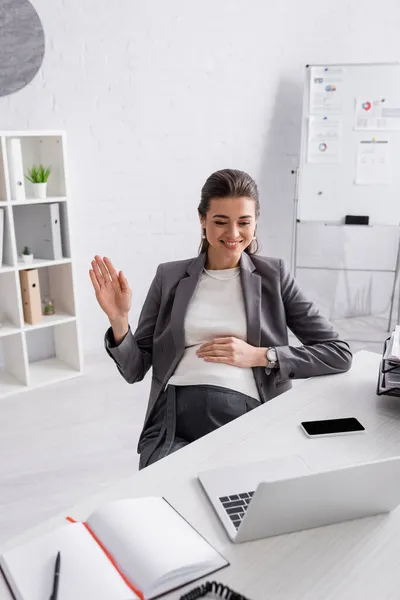 Joyeuse femme d'affaires enceinte agitant la main lors d'un appel vidéo — Photo de stock