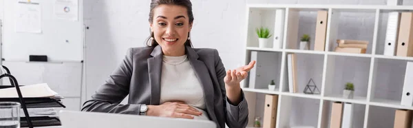 Allegra donna d'affari incinta gesticolando vicino al computer portatile in ufficio, banner — Foto stock