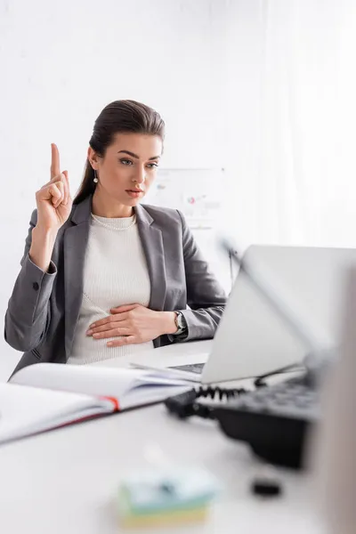 Young pregnant businesswoman pointing up and looking at laptop in office — Stock Photo