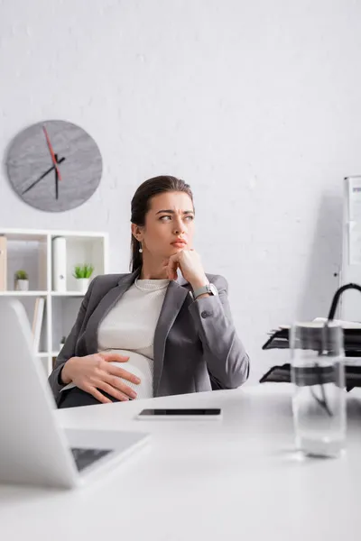 Pensativo embarazada mujer de negocios mirando lejos cerca de gadgets en escritorio - foto de stock