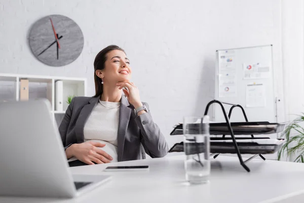 Feliz embarazada mujer de negocios mirando lejos cerca de gadgets en el escritorio - foto de stock