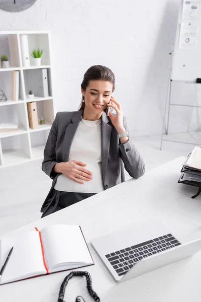 Felice donna d'affari incinta che parla su smartphone vicino al computer portatile sulla scrivania — Foto stock