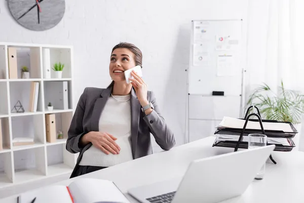 Cheerful pregnant businesswoman talking on smartphone near laptop on desk — Stock Photo