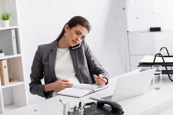 Joven mujer de negocios embarazada hablando en el teléfono inteligente y la escritura en el portátil cerca de la computadora portátil en el escritorio - foto de stock