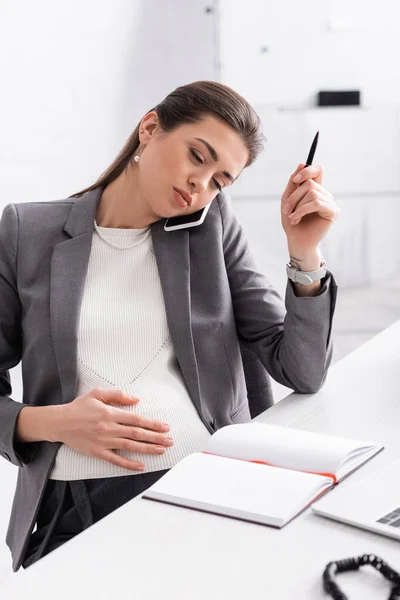 Joven embarazada mujer de negocios hablando en el teléfono inteligente y la celebración de la pluma cerca del ordenador portátil en el escritorio - foto de stock