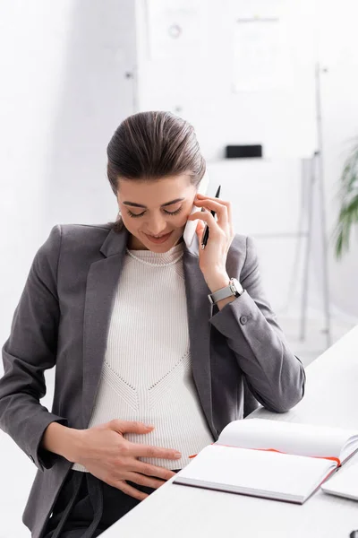 Счастливая беременная деловая женщина разговаривает на смартфоне и трогает живот — стоковое фото