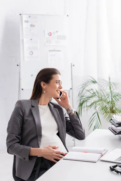 Alegre embarazada mujer de negocios hablando en smartphone en la oficina - foto de stock