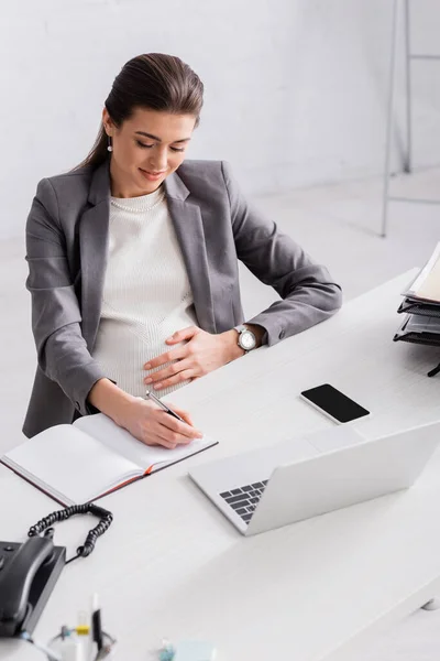 Pregnant businesswoman writing in notebook near gadgets on desk — Stock Photo