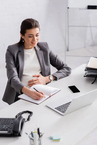 Sonriente embarazada mujer de negocios escribiendo en cuaderno cerca de gadgets en el escritorio - foto de stock