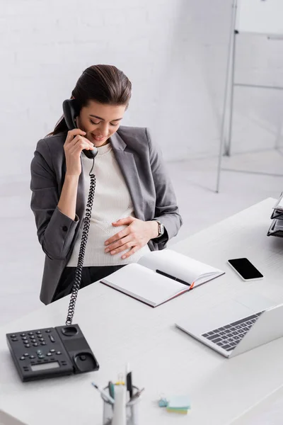 Весела вагітна бізнес-леді розмовляє на ретро телефоні біля гаджетів — стокове фото