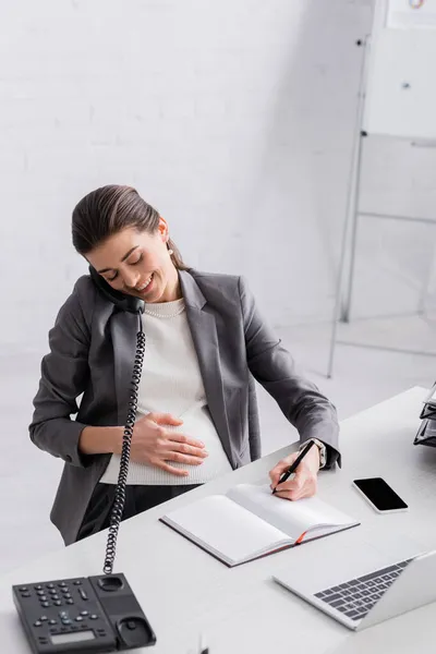 Cheerful pregnant businesswoman talking on retro telephone and writing in notebook — Stock Photo