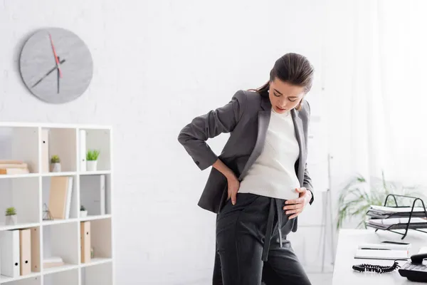 Exhausted pregnant woman touching back near desk in office — Stock Photo