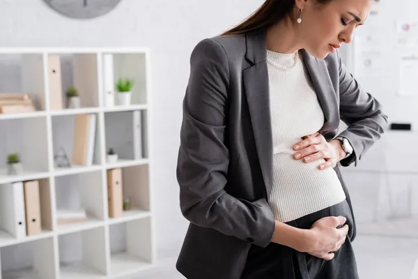 Morena embarazada mujer de negocios tocando vientre mientras se siente calambre en la oficina - foto de stock