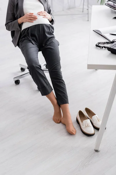 Vista cortada de mulher grávida sem sapatos sentada na cadeira no escritório — Fotografia de Stock
