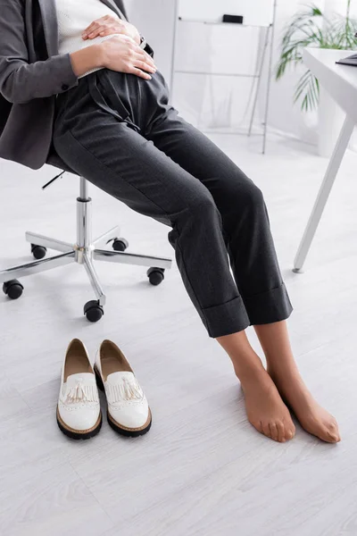 Vista ritagliata di donna incinta senza scarpe seduta sulla sedia vicino alla scrivania — Foto stock