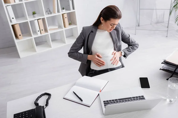 Blick aus der Vogelperspektive auf unzufriedene schwangere Geschäftsfrau, die Bauch berührt, während sie im Büro Krämpfe verspürt — Stockfoto