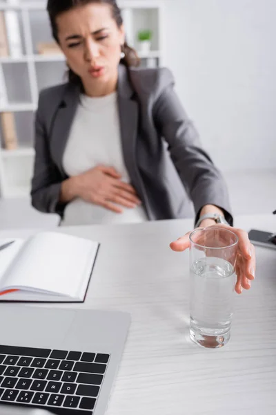 Femme d'affaires enceinte floue se sentant crampe et atteignant un verre d'eau sur la table de bureau — Photo de stock