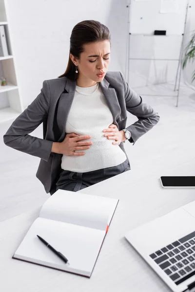 Femme d'affaires enceinte toucher le ventre tout en se sentant crampes près de gadgets sur le bureau — Photo de stock