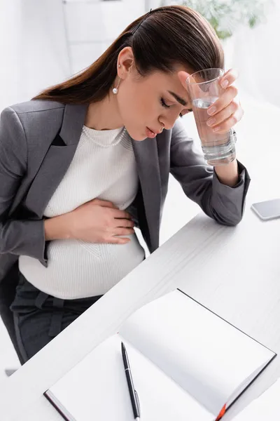 Donna d'affari incinta che tiene un bicchiere d'acqua mentre si sente male — Foto stock