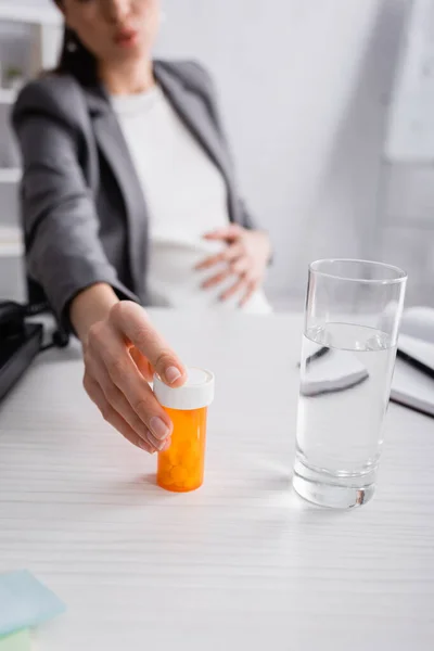 Ausgeschnittene Ansicht einer verschwommenen Schwangeren, die mit Tabletten in der Nähe eines Wasserglases auf dem Schreibtisch steht — Stockfoto