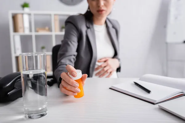 Обрезанный вид беременной женщины, достигающей банка с таблетками возле стакана воды на столе — стоковое фото