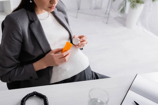 Vista recortada de la mujer embarazada sosteniendo frasco con pastillas en la oficina - foto de stock