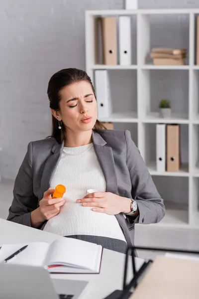 Mulher grávida segurando jar com pílulas enquanto se sente cãibra no escritório — Fotografia de Stock