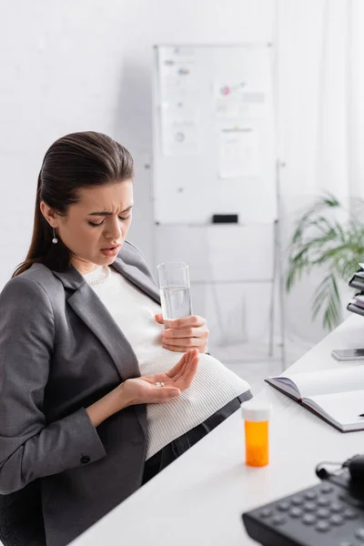 Mulher grávida segurando pílulas e vidro de água enquanto se sente cãibra no escritório — Fotografia de Stock