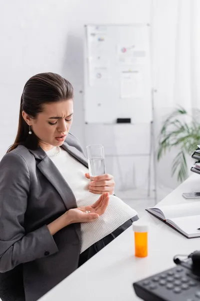 Pregnant woman holding pills and glass of water while feeling cramp in office — Stock Photo